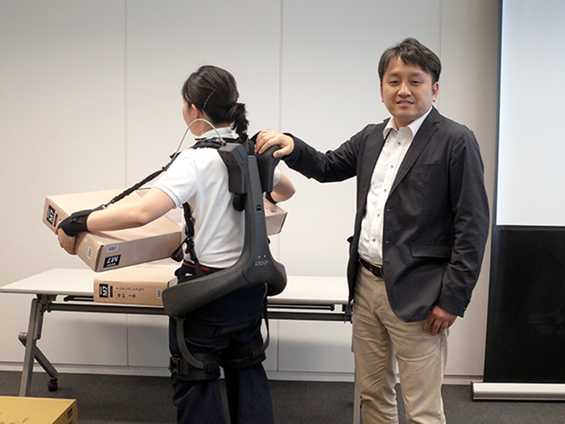 ATOUN 代表取締役社長の藤本弘道氏（右）と腕の補助機能がついたパワーアシストスーツ「ATOUN MODEL Y」