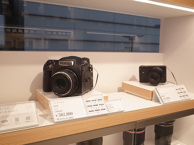 　店内の奥に進むとペンタックス「645Z」など中判デジタル一眼レフカメラが展示。ガラスケースには入っておらず、そのまま手にとれる。

　カメラはすべて専用の什器に陳列されており、プライスカードや機能表示なども白黒のシンプルな表示で統一。カメラ台も独自のものを採用することで、スッキリとした印象になるほか、コンパクトなスペースでも多くのカメラを展示できるとのこと。1つの棚の幅は約70cmになっており、厚みのある棚板を採用することで、没入感があり、カメラに集中しやすい環境にしているという。