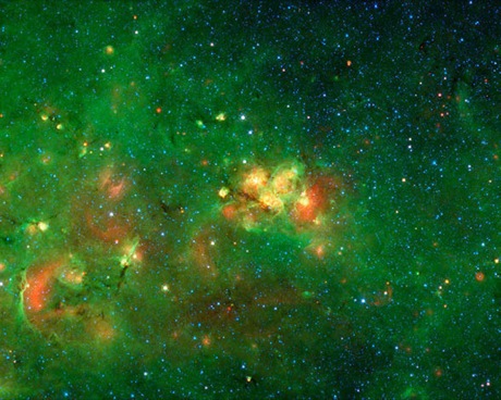 　この画像の中心に見えている黄色がかった赤色の星雲はたて座にあり、ちりの雲に隠れていることから一般的な名称が付けられていない。この暗いベールを透かして、この素晴らしい隠された星雲を発見するには、可視光よりも長い波長を観測する、Spitzer宇宙望遠鏡のような赤外線望遠鏡が必要だ。