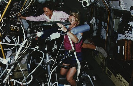 　自転車エルゴメータを使う宇宙飛行士のRhea Seddon氏。宇宙飛行士のTammy Jernigan氏が実験のモニタリングを手伝っている。