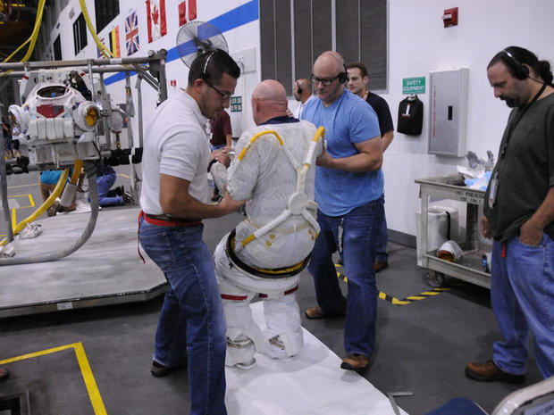 　Scott Kelly氏が宇宙服のズボンを着用し始めた後、同氏が立ち上がるのを助けるNBLの技術者たち。