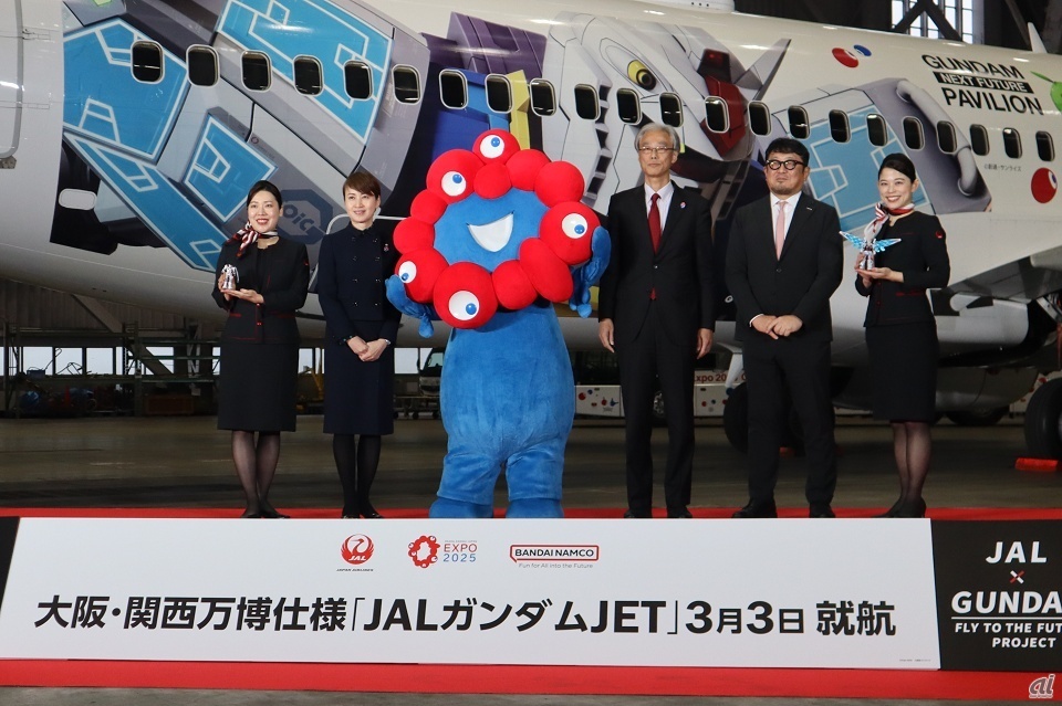 特別仕様のガンプラを持つ客室乗務員に挟まれた、（左から）日本航空 常務執行役員 西日本支店長 宮坂久美子氏、公式キャラクター「ミャクミャク」、バンダイナムコホールディングス 取締役 桃井信彦氏、2025年日本国際博覧会協会 副事務総長 高科淳氏
